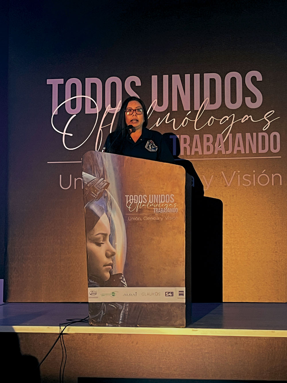 Doctora oftalmóloga hablando desde el podio del escenario principal del evento Todos unidos: oftalmólogos trabajando