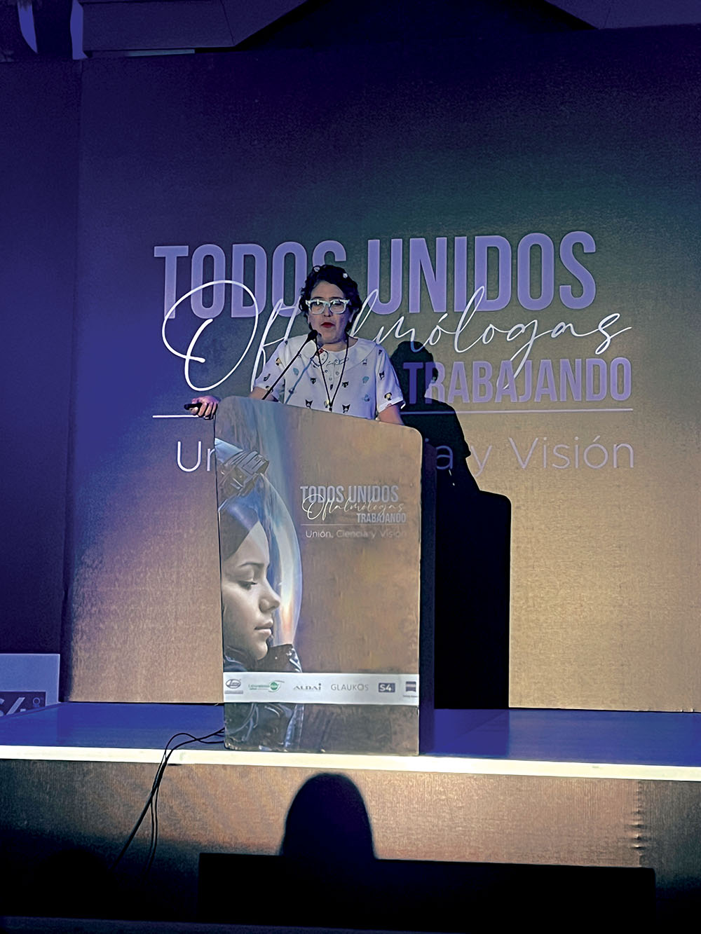 Doctora oftalmóloga hablando desde el podio del escenario principal del evento Todos unidos: oftalmólogos trabajando
