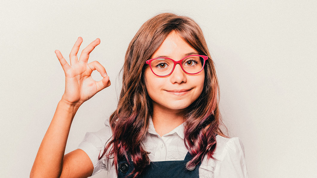 Niña de aproximadamente 10 años, usando lentes ópticos color rojo, mientras sonríe a la cámara y hace una señal de "OK" con la mano