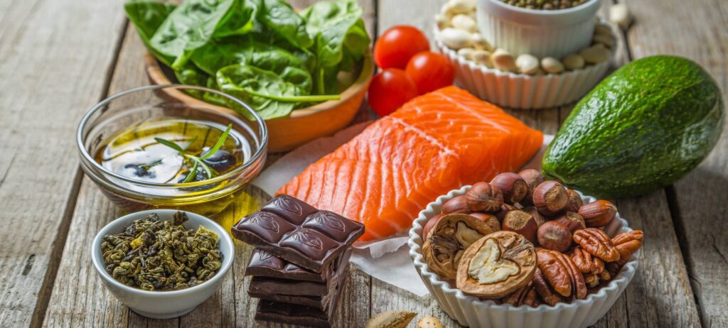 Alimentos variados que aportan nutrición para los ojos (salmón, aguacate, aceite de oliva, nueces, espinacas, chocolate y otros) acomodados sobre una mesa de madera