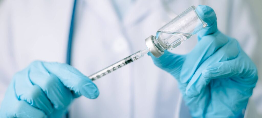 Manos de un médico usando guantes de latex azul sosteniendo con la mano derecha una jeringa y con la mano izquierda una ampolleta que contiene medicamento para enfermedad de los ojos