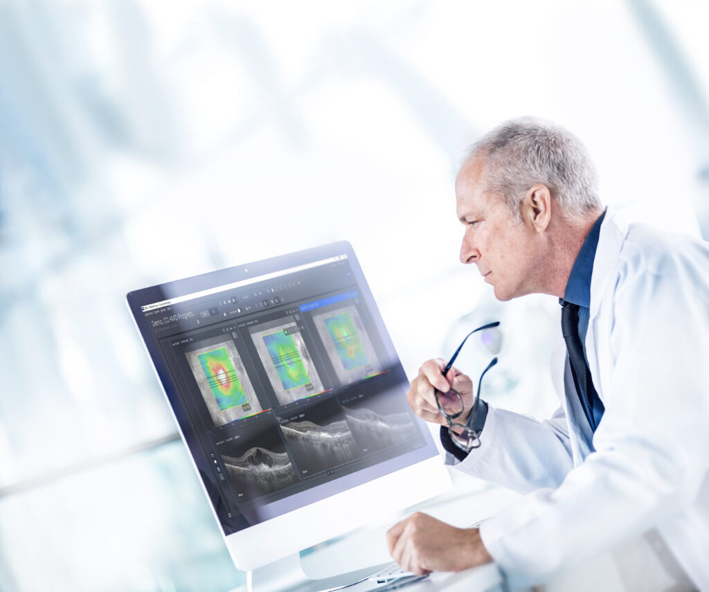 Médico en bata blanca observa atentamente imágenes clínicas de retina en una pantalla de computadora, que incluyen escaneos OCT y mapas de densidad visual, en un entorno moderno y luminoso.